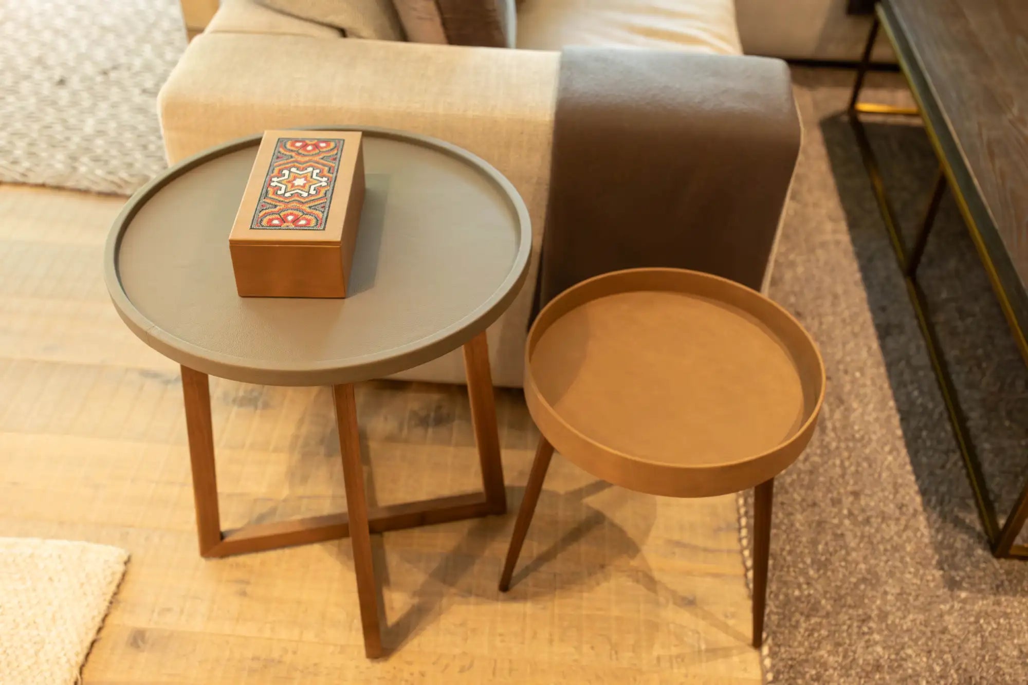 La imagen muestra dos mesas laterales redondas con patas de madera y superficies superiores planas, una con un acabado gris apagado y la otra en un tono marrón cálido.