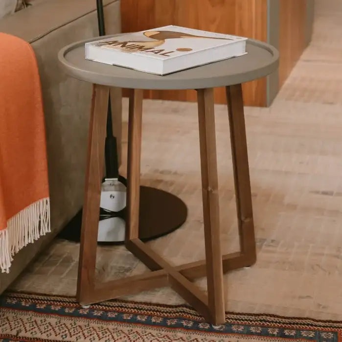 Mesa lateral circular gris con un diseño elegante y minimalista que presenta cuatro patas de madera anguladas que se intersectan en la base.