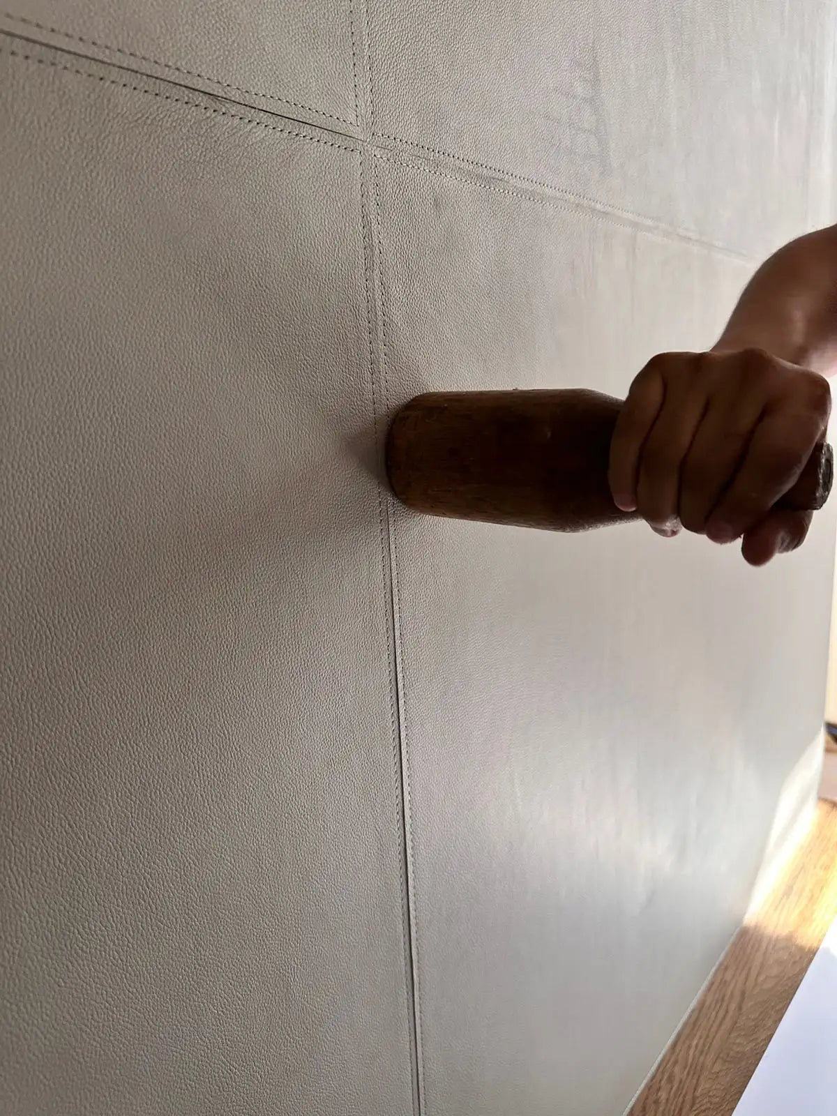 Una mano agarra un mazo de madera marrón oscuro mientras golpea la costura de una superficie de cuero gris claro.