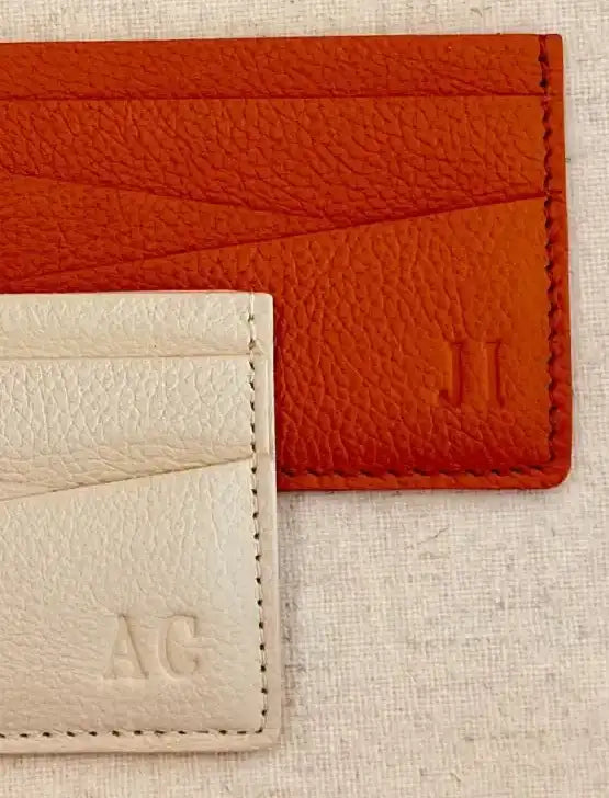 Dos portatarjetas de cuero, uno en naranja vibrante y el otro en blanco cremoso, ambos con iniciales en relieve.